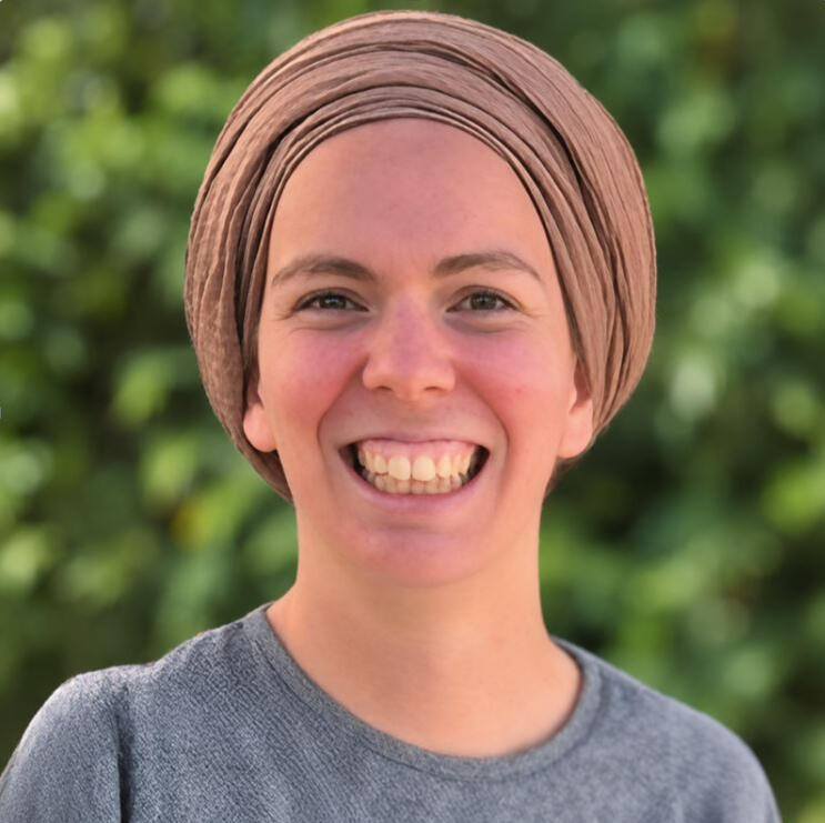 Portrait of Yemima Shindler, smiling and wearing a beige headscarf against a leafy green background.